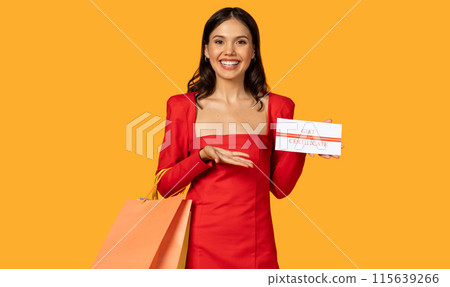 A cheerful woman stands against a vibrant orange background, elegantly dressed in a red dress and holding a gift certificate in one hand with several shopping bags draped over her arm. A cheerful woman stands against a vibrant orange background, elegantly dressed in a red dress and holding a gift certificate in one hand with several shopping bags draped over her arm. 115639266