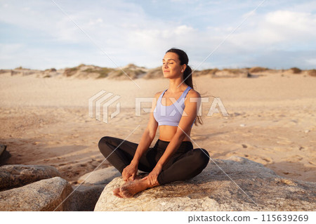 Happy calm young caucasian lady enjoy workout, breathing exercises, practice yoga in lotus position, meditation in morning on sea beach. Hobby outdoor, sports, lifestyle and health care Happy calm young caucasian lady enjoy workout, breathing exercises, practice yoga in lotus position, meditation in morning on sea beach. Hobby outdoor, sports, lifestyle and health care 115639269