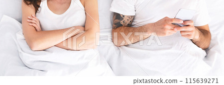 A woman with her arms crossed lies in a bed beside a man who is using his phone. She appears upset or disinterested, while the man is engrossed in the screen, cropped, panorama A woman with her arms crossed lies in a bed beside a man who is using his phone. She appears upset or disinterested, while the man is engrossed in the screen, cropped, panorama 115639271