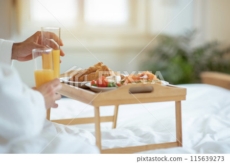 Romantic Breakfast In Bed. Closeup Of Tray Filled With Delicious Eats, Laid On Bed For A Couple, Hands Grasping Glasses Of Fresh Orange Juice At Modern Hotel Bedroom Interior. Cropped 115639273