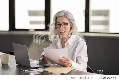 Business, Employment. Excited Human Resources Manager Lady Reading Resumes And Job Applications, Enjoying Work And Having Fun At Workplace, Sitting At Laptop And Holding Paper CVs Indoors 115639274