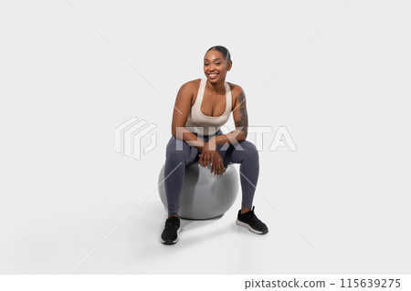 African American young woman with short black hair is sitting on an exercise ball in a studio setting. She is wearing a white tank top and grey leggings 115639275