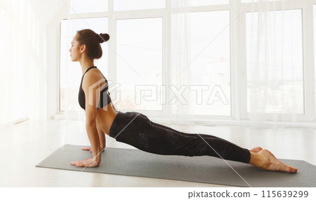A woman is engaged in a yoga pose on a yoga mat. She is focused and balanced, demonstrating flexibility and strength in her body. The background is minimal, emphasizing her form and concentration. A woman is engaged in a yoga pose on a yoga mat. She is focused and balanced, demonstrating flexibility and strength in her body. The background is minimal, emphasizing her form and concentration. 115639299