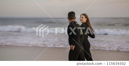 Happy Smiling Surfers Couple Running Together Along The Beach, Romantic Young Man And Woman Wearing Wetsuits Surfing Together On Ocean Shore, Enjoying Water Sports, Panorama With Copy Space 115639302