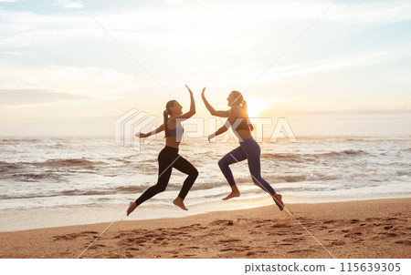 Cheerful young caucasian women enjoy morning workout, have fun, jump, freeze in air, give high five on sea beach. Great sports result, lifestyle and body care, weight loss outdoor together 115639305