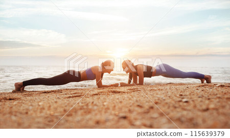 Cheerful young caucasian women enjoy morning workout, doing plank exercise on sea beach, panorama, full length. Body care together, weight loss, sports and fit lifestyle outdoor 115639379