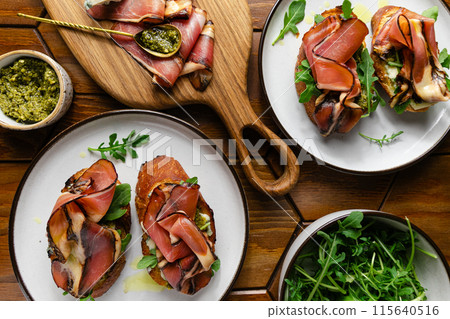 Appetizer toasts with prosciutto, pesto sauce, cheese and arugula on a table on a table Appetizer toasts with prosciutto, pesto sauce, cheese and arugula on a table on a table 115640516