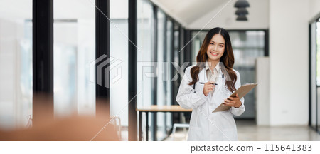A woman in a white lab coat is standing in a room with a clipboard in her hand. She is a doctor or a nurse, and she is smiling. The room is well-lit and has a modern, clean appearance 115641383