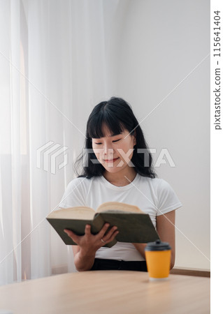 A young woman in casual clothing reading a book at home with a cup of coffee on a wooden table, enjoying a peaceful and relaxed atmosphere. A young woman in casual clothing reading a book at home with a cup of coffee on a wooden table, enjoying a peaceful and relaxed atmosphere. 115641404