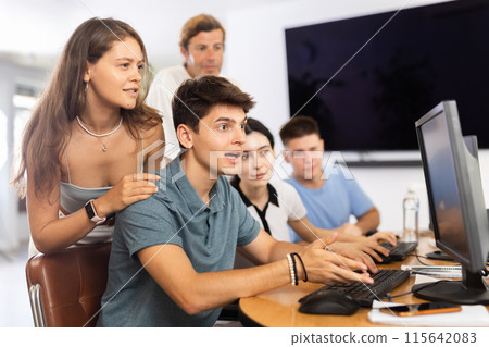 Girl stands behind sitting students, looks at monitor and helps in execution of programing teamwork Girl stands behind sitting students, looks at monitor and helps in execution of programing teamwork 115642083