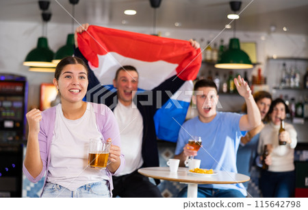 Company of excited young adult sports fans waving Dutch flag and supporting national team with beer in pub 115642798