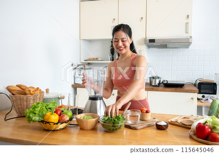 A beautiful, healthy Asian woman in sportswear is making her healthy smoothie drink in the kitchen. A beautiful, healthy Asian woman in sportswear is making her healthy smoothie drink in the kitchen. 115645046