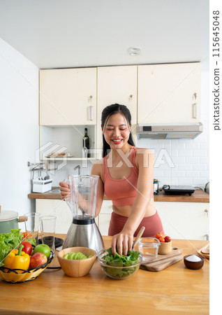 A beautiful, healthy Asian woman in sportswear is making her healthy smoothie drink in the kitchen. A beautiful, healthy Asian woman in sportswear is making her healthy smoothie drink in the kitchen. 115645048