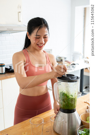 A strong, healthy Asian woman in sportswear is making a green smoothie with a blender in the kitchen A strong, healthy Asian woman in sportswear is making a green smoothie with a blender in the kitchen 115645062