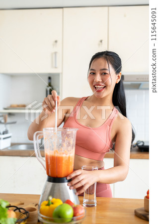 A fit Asian woman in sportswear making a smoothie or healthy juice in the kitchen with a blender. A fit Asian woman in sportswear making a smoothie or healthy juice in the kitchen with a blender. 115645091