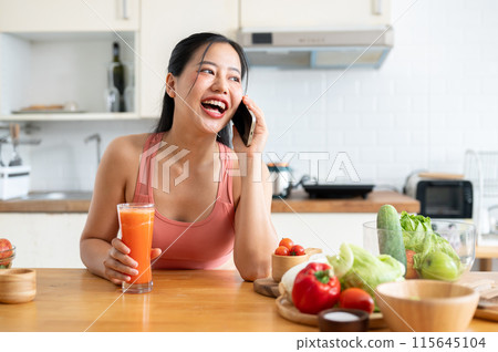 A charming Asian woman in sportswear is talking on the phone in the kitchen while drinking smoothie. 115645104