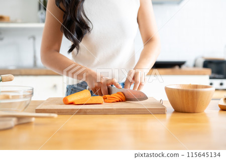 A woman slicing carrots on a cutting board at a wooden kitchen tabletop, cooking in the kitchen. A woman slicing carrots on a cutting board at a wooden kitchen tabletop, cooking in the kitchen. 115645134