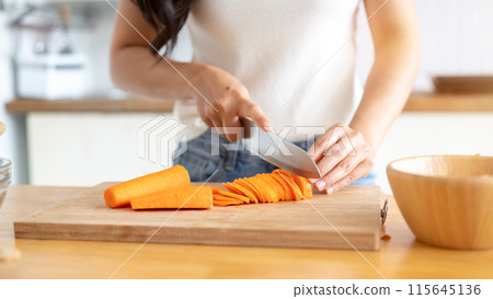 A woman slicing carrots on a cutting board at a wooden kitchen tabletop, cooking in the kitchen. 115645136