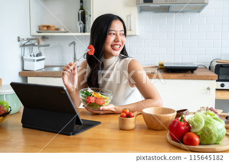 An attractive Asian woman eating salad while working on her digital tablet in the kitchen. An attractive Asian woman eating salad while working on her digital tablet in the kitchen. 115645182