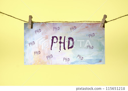 The word PHD is written on paper suspended from a rope on a yellow background The word PHD is written on paper suspended from a rope on a yellow background 115651218