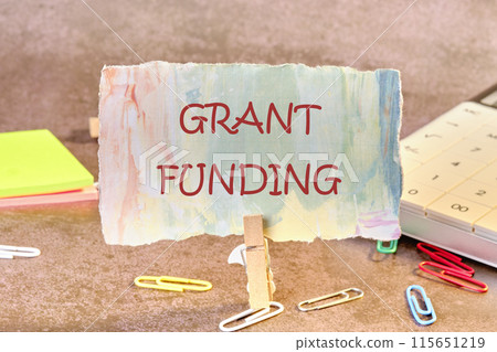 Business and grant funding concept. Copy space. Words GRANT FUNDING on paper in a clothespin in a composition with a calculator, stickers and scattered office paper clips Business and grant funding concept. Copy space. Words GRANT FUNDING on paper in a clothespin in a composition with a calculator, stickers and scattered office paper clips 115651219