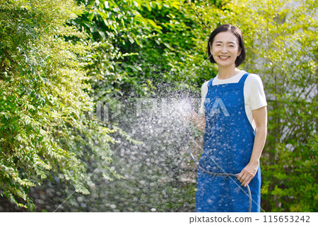 微笑的中年婦女在花園裡的綠樹澆水 微笑的中年婦女在花園裡的綠樹澆水 115653242