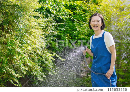 微笑的中年婦女在花園裡的綠樹澆水 115653356