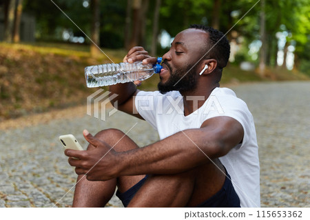 Young adult man resting on a park after a run with a bottle of water and smartphone. Outdoor workout Young adult man resting on a park after a run with a bottle of water and smartphone. Outdoor workout 115653362