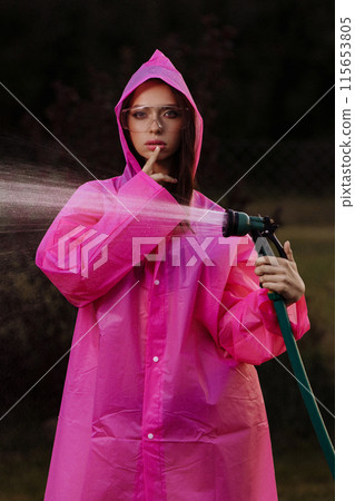 A glamour woman in a pink raincoat is spraying water from a hose 115653805
