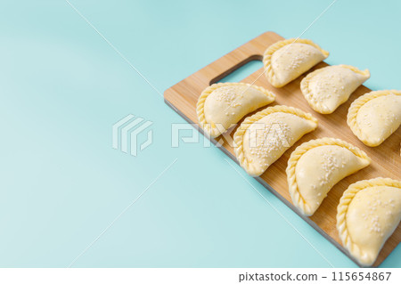 Raw samosas with sesame on a wooden on board on blue table ready for baking. Traditional Central Asian cuisine, frozen semi-finished products, copy space. 115654867