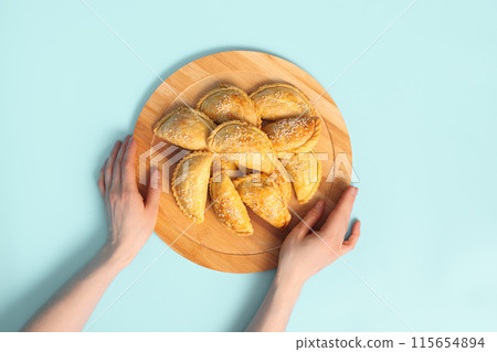 Top view of Woman Holding Wooden Plate with Traditional Central Asian Samsa Pies on Blue Background. Top view of Woman Holding Wooden Plate with Traditional Central Asian Samsa Pies on Blue Background. 115654894