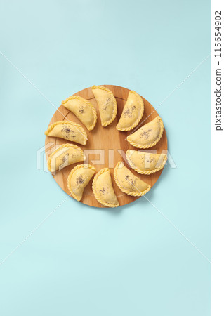 Flat lay on raw samsa on a wooden round board on blue background ready for baking. Traditional Central Asian cuisine bakery, frozen semi-finished products, copy space. 115654902