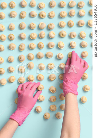 A chef's hands in a rubber glove carefully lays out semi-cooked raw dumplings on a blue background, top view. A chef's hands in a rubber glove carefully lays out semi-cooked raw dumplings on a blue background, top view. 115654910