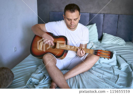 a young man is learning to play the guitar at home, a cat is lying next to him and listening a young man is learning to play the guitar at home, a cat is lying next to him and listening 115655559