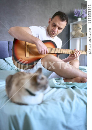 a young man is learning to play the guitar at home, a cat is lying next to him and listening a young man is learning to play the guitar at home, a cat is lying next to him and listening 115655561