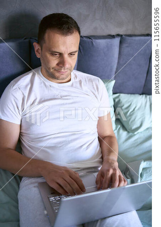 a young man is working on a laptop and a smartphone in bed, a cat is lying next to him 115655596
