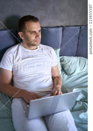 a young man is working on a laptop and a smartphone in bed, a cat is lying next to him a young man is working on a laptop and a smartphone in bed, a cat is lying next to him 115655597