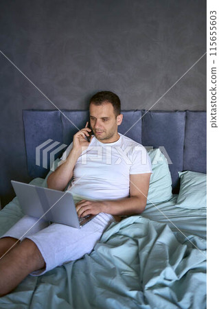 a young man on the bed working on a laptop and talking on the phone a young man on the bed working on a laptop and talking on the phone 115655603