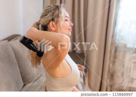 Girl at home holding a therapeutic percussion vibrating massage gun in her hand and massaging her back muscles Sports recovery concept after a workout. Girl at home holding a therapeutic percussion vibrating massage gun in her hand and massaging her back muscles Sports recovery concept after a workout. 115660997