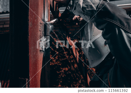 A welder welds metal hinges on a gate while wearing a protective mask. During welding, sparks fly and bright flashes are visible. A man repairs a gate by welding new hinges. 115661392