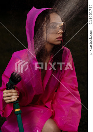 A glamour woman in a pink raincoat is spraying water from a hose 115661753