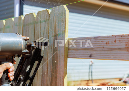 Worker uses an air pneumatic gun to nail wooden planks to fence during work Worker uses an air pneumatic gun to nail wooden planks to fence during work 115664057