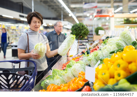 Elderly woman examines china cabbage in vegetable section to choose juiciest cabbage Elderly woman examines china cabbage in vegetable section to choose juiciest cabbage 115664457