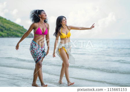 Two women are walking on the beach, one wearing a pink bikini 115665153