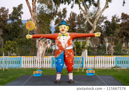 Joyful statue of a clown with outstretched arms from which a swing hangs in the park Joyful statue of a clown with outstretched arms from which a swing hangs in the park 115667576