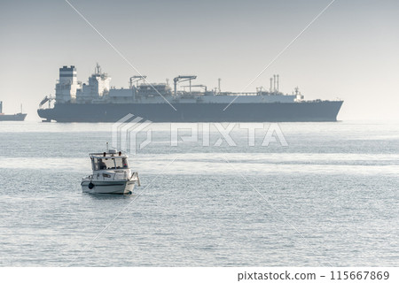 Small boat sailing near a large container ship on a hazy day at Akrotiri Bay, Cyprus 115667869