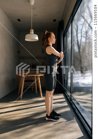 Full length portrait of sporty woman preforming warm-up exercise, stretching arm muscles before workout indoors, standing by window with beautiful snowy winter nature view on sunny day, looking away. Full length portrait of sporty woman preforming warm-up exercise, stretching arm muscles before workout indoors, standing by window with beautiful snowy winter nature view on sunny day, looking away. 115669584