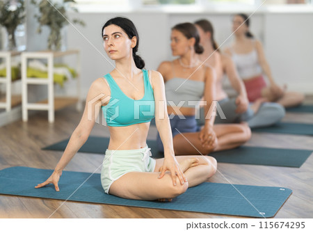 Women maintaining mental and physical health attending group yoga class at modern studio, practicing twisting pose Women maintaining mental and physical health attending group yoga class at modern studio, practicing twisting pose 115674295