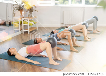 Together with students, she lies on mat, holds fitball between knees and raises and lowers buttocks 115674716