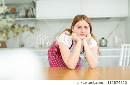 Calm positive teen girl wearing red polka dot apron and leaning on kitchen countertop in cuisine 115674850